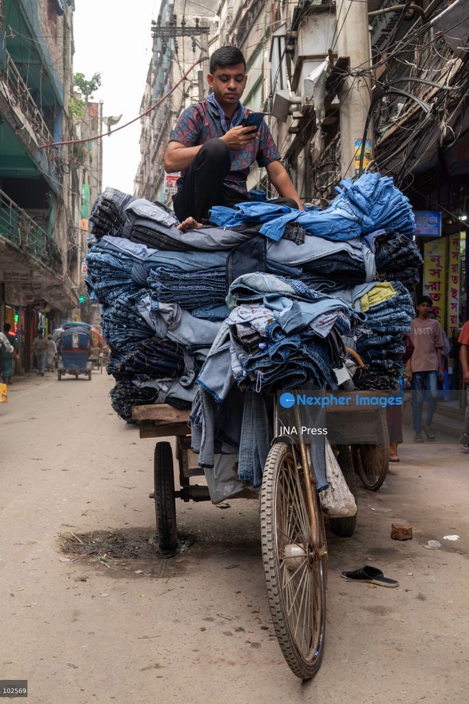 KALIGANJ, DHAKA, BANGLADESH, OCTOBER 7, 2026: Garment traders and workers transport large bundles of denim clothing through a busy market in Kaliganj, Dhaka. Jeans and other garments are stacked outside shops and moved through crowded streets by handcart, bicycle and truck for distribution and sale. The market forms part of the local apparel trade, where workers and traders handle large volumes of clothing moving between wholesalers, retailers and buyers. Photo by Bipul Ahmed / JNA Press. (Photo by Bipul Ahmed/JNA Press via Nexpher Images)